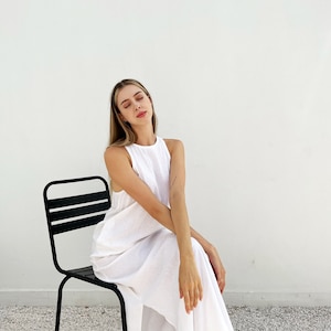 Puede incluir: Una mujer con un vestido blanco de lino se sienta en una silla de metal negro contra una pared blanca.