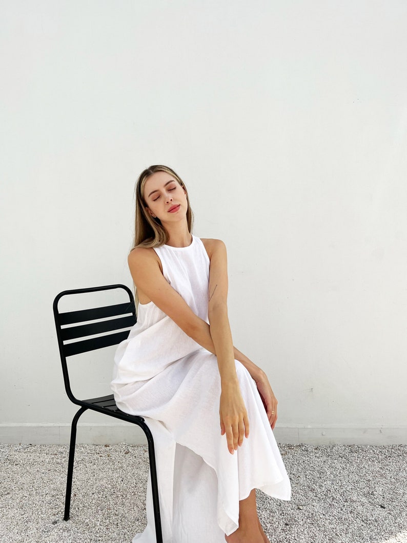 Puede incluir: Una mujer con un vestido blanco de lino se sienta en una silla de metal negro contra una pared blanca.