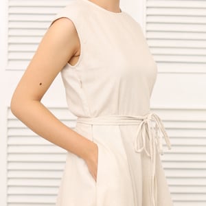 May include: A sleeveless, light beige linen dress with a tie belt and pockets. The dress has a simple, classic design.