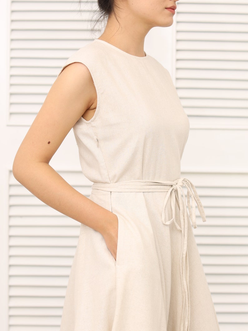 May include: A sleeveless, light beige linen dress with a tie belt and pockets. The dress has a simple, classic design.