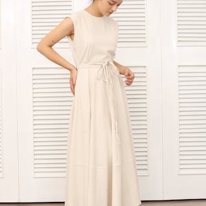 May include: A beige linen maxi dress with a tie waist and a flowy skirt. The dress is sleeveless and has a round neckline.