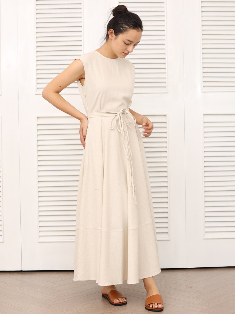 May include: A beige linen maxi dress with a tie waist and a flowy skirt. The dress is sleeveless and has a round neckline.