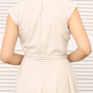 May include: A beige linen dress with a V-neckline and short sleeves. The dress has a tie belt at the waist and a flowy skirt.