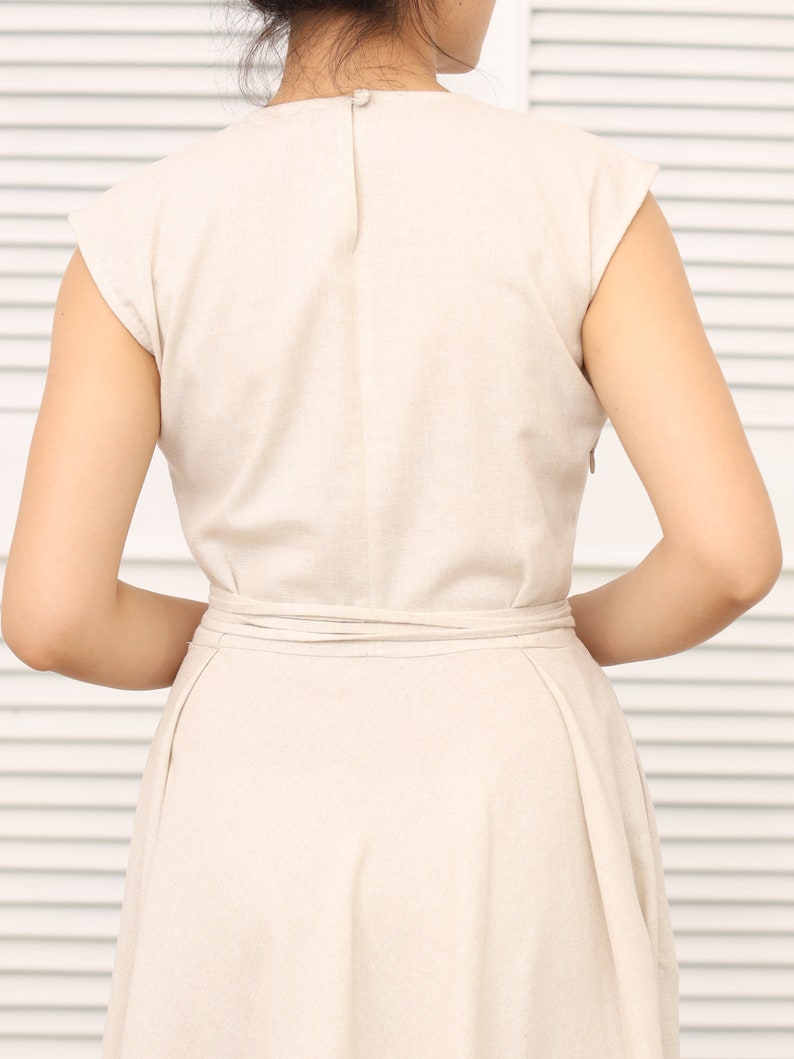 May include: A beige linen dress with a V-neckline and short sleeves. The dress has a tie belt at the waist and a flowy skirt.