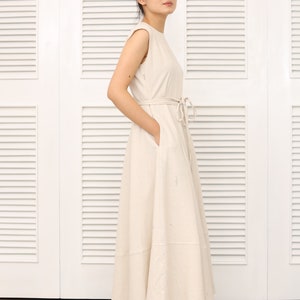 May include: A woman wearing a light beige sleeveless maxi dress with a tie at the waist. The dress has a slightly flared skirt and is made of a lightweight fabric.