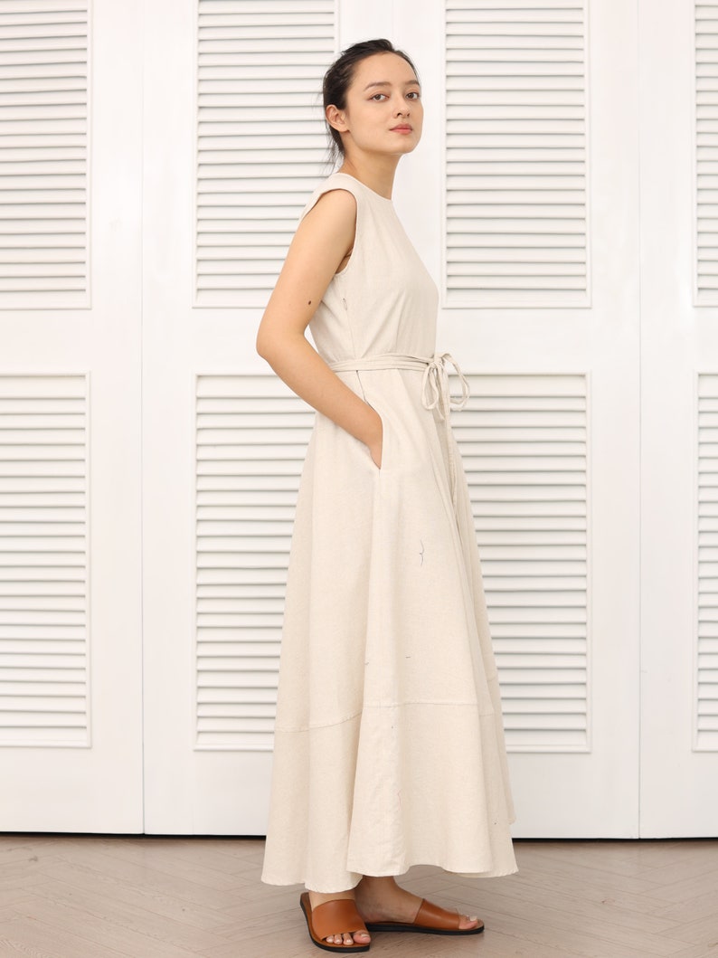 May include: A woman wearing a light beige sleeveless maxi dress with a tie at the waist. The dress has a slightly flared skirt and is made of a lightweight fabric.