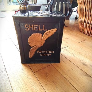 Puede incluir: Bidón negro vintage de Shell Aviation Spirit con logotipo y letras doradas. El bidón tiene un asa y un pequeño pico. Las palabras "SHELL" y "AVIATION SPIRIT" son visibles.