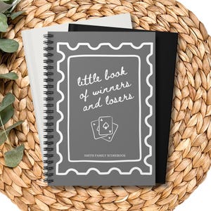 May include: A spiral-bound notebook with a gray cover, featuring the text "little book of winners and losers" and a playing card graphic. The notebook is on a woven, circular placemat with eucalyptus sprigs.