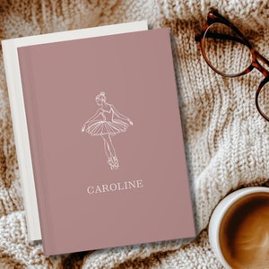 May include: A dusty rose journal with a white line drawing of a ballerina and the name "CAROLINE" in a serif font. The journal is on a cream-colored knitted blanket, with a pair of tortoiseshell glasses and a cup of coffee.