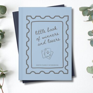 K&ouml;nnte beinhalten: Ein hellblaues Buch mit dem Text "little book of winners and losers" und zwei Spielkarten. Das Buch hat ein graues Rahmendesign und die Worte "SMITH FAMILY SCOREBOOK" unten. Eukalyptuszweige sind im Hintergrund.