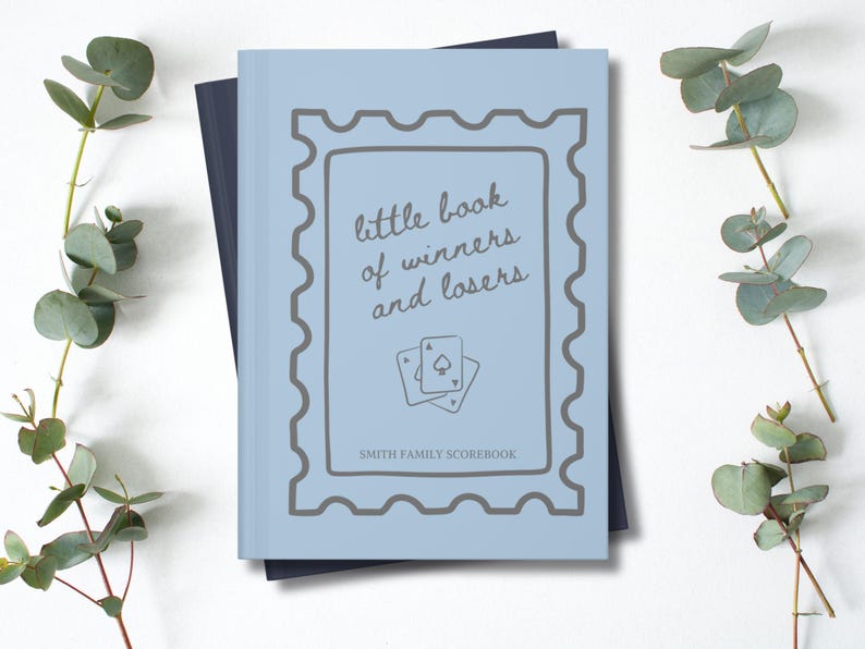 K&ouml;nnte beinhalten: Ein hellblaues Buch mit dem Text "little book of winners and losers" und zwei Spielkarten. Das Buch hat ein graues Rahmendesign und die Worte "SMITH FAMILY SCOREBOOK" unten. Eukalyptuszweige sind im Hintergrund.