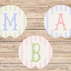 May include: Three round car coasters with a striped background and a single initial in the center. The initials are M, A, and B, in blue, pink, and green, respectively. The coasters are white with pastel stripes.