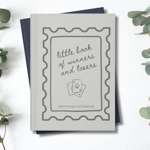 K&ouml;nnte beinhalten: Ein hellgraues Buch mit dem Text "little book of winners and losers" und zwei Spielkarten. Das Buch ist mit einem grauen Rahmen versehen und liegt auf einem dunkelblauen Buch. Eukalyptuszweige befinden sich an den Seiten.