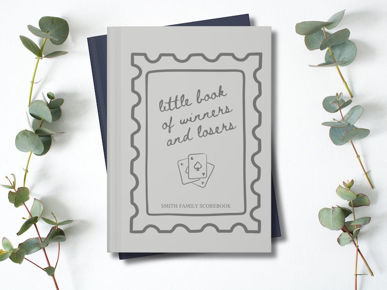 K&ouml;nnte beinhalten: Ein hellgraues Buch mit dem Text "little book of winners and losers" und zwei Spielkarten. Das Buch ist mit einem grauen Rahmen versehen und liegt auf einem dunkelblauen Buch. Eukalyptuszweige befinden sich an den Seiten.