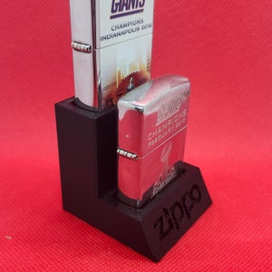 May include: Two silver Zippo lighters with black and white graphics. The top lighter has the text "Giants Champions Indianapolis 2012". The bottom lighter has the text "Champions February 4, 2012". Both lighters are in a black plastic stand with the word "Zippo" printed on the front.