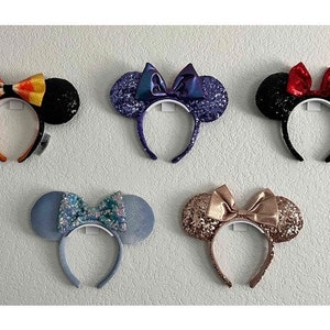 Pode incluir: Cinco pares de orelhas de Minnie Mouse com lantejoulas e laços de diferentes cores. As orelhas são pretas, azuis, douradas, roxas e vermelhas.