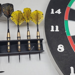 Może przedstawiać: Zestaw sześciu czarnych i złotych lotków z żółtymi i czarnymi wzorzystymi lotkami. Lotki znajdują się w czarnym plastikowym uchwycie. W tle znajduje się czarno-biała tarcza do lotków z czerwonymi i zielonymi sekcjami. Na tarczy do lotków widoczne są liczby 8, 11 i 14.