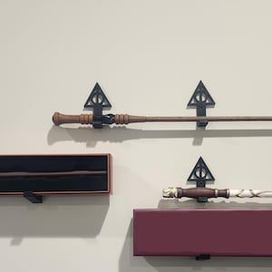 May include: Three magic wands are displayed on a wall. One wand is in a black box, and the other two are mounted on black brackets. The wands are brown, white, and gold, with intricate designs.