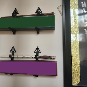 May include: Two wooden wands are displayed on shelves with black triangle-shaped brackets. The shelves are mounted on a white wall. The wands are resting on a green and purple shelf. The text "HARRY POTTER" is visible on a black framed sign with gold glitter.
