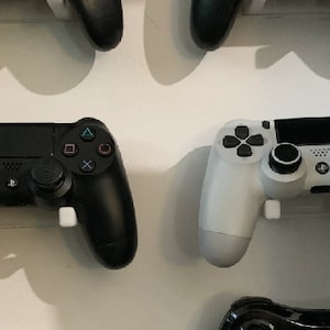 Puede incluir: Dos mandos de videojuegos montados en una pared blanca. El mando de la izquierda es negro con botones blancos y el mando de la derecha es blanco con botones negros.