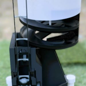 Puede incluir: Un dispositivo mecánico en blanco y negro con una parte superior e inferior blancas y un diseño en espiral. El dispositivo tiene un asa y está colocado sobre una superficie verde. El dispositivo está diseñado para dispensar una bola.