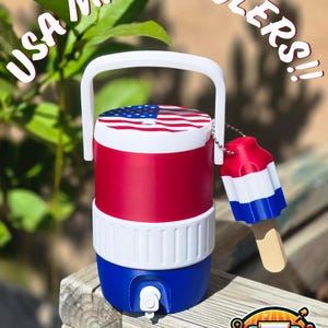 Peut inclure: Une mini glacière rouge, blanche et bleue avec une poignée, ornée d'un drapeau américain sur le dessus. La glacière est accompagnée d'un porte-clés en forme de glace. Le texte "USA MINI COOLERS!!" est arqué au-dessus.