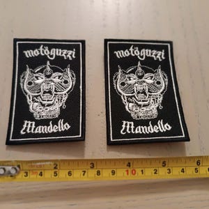May include: Two rectangular black embroidered patches featuring white thread designs of a stylized eagle head and the text motoguzzi and Mandello. A yellow measuring tape is positioned below the patches, showing a width of approximately 7.6 cm.