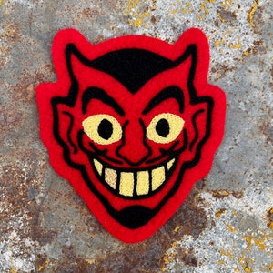 May include: Red felt devil face patch with black outline and yellow eyes. The devil is smiling with a row of yellow teeth.