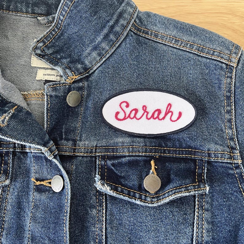 May include: A blue denim jacket with a white oval patch that says "Sarah" in red cursive lettering.