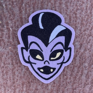 May include: Purple felt patch with an embroidered cartoon face of a vampire with black hair, yellow eyes, and a toothy grin.