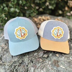 May include: Two blue and gray trucker hats with a white mesh back. Both hats have a yellow, orange, and green embroidered patch with the letters "SLO" in a circle.