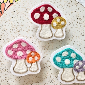 May include: Three embroidered felt mushroom patches. One is red with white dots, one is pink with white dots, and one is teal with white dots. Each mushroom has a smaller mushroom on top.