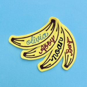 May include: Four yellow banana-shaped felt patches with dark brown embroidered outlines. Each patch features a name stitched in cursive thread: Olivia in blue, Abby in dark pink, Noah in black, and Benya in dark red, arranged in a fan pattern.