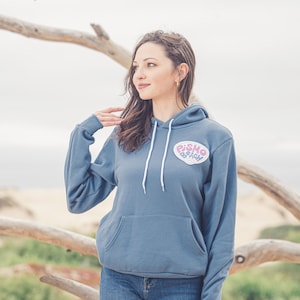 May include: A woman wearing a blue hooded sweatshirt with a pink and white heart-shaped patch that says "Pismo Beach".