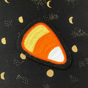 May include: Embroidered candy corn patch with yellow, orange, and white sections, outlined in black. The patch is on a black fabric with gold crescent moons and starbursts, perfect for Halloween or fall crafts.