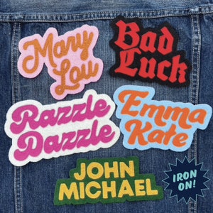 May include: A collection of iron-on patches featuring various names and phrases, including "Mary Lou", "Bad Luck", "Razzle Dazzle", "Emma Kate", "John Michael", and "Iron On!" The patches are made of felt and have a variety of colors, including pink, orange, red, blue, green, and yellow.