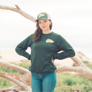 May include: A woman wearing a dark green sweatshirt with a yellow and orange "Slo" patch on the front. She is also wearing a green and white baseball cap with the word "Slo" on it. The woman is standing in front of a wooden branch.