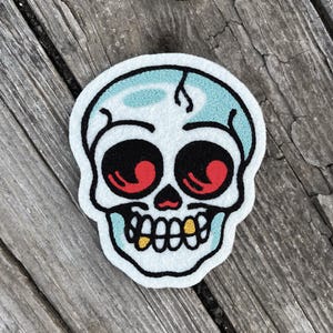 May include: A white skull-shaped patch with black, red, and gold accents. The skull has large black eye sockets with red details, a turquoise top, and gold teeth. The patch is on a wooden surface.