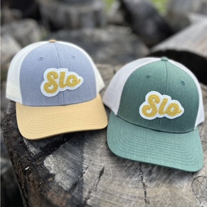 May include: Two trucker hats with a white mesh back and a gray and green front. Both hats have a yellow felt patch with the word "Slo" in a retro font.