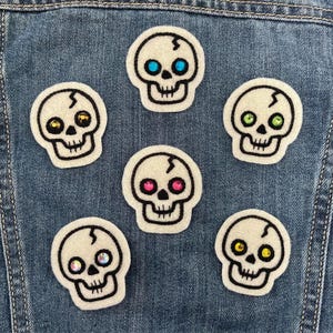 May include: Six white felt skull patches with black embroidered outlines and colorful rhinestone eyes, including blue, yellow, green, pink, and iridescent gems, arranged on a blue denim fabric background.