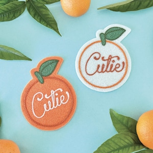May include: Two embroidered felt orange patches with the word "Cutie" written in white cursive. One patch is orange and the other is white with an orange outline.