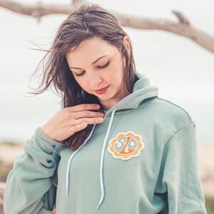 May include: A sage green hooded sweatshirt with a white drawstring and a circular patch featuring the letters "SLO" in a retro font. The patch is orange and yellow with a white outline.