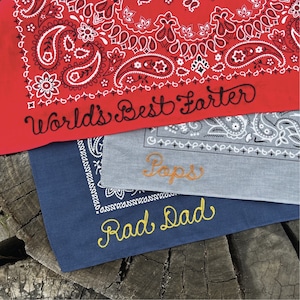 May include: A collection of bandanas in red, gray, and navy blue, each with a unique embroidered message. The red bandana features the text "World's Best Farter", the gray one says "Pops", and the navy blue one reads "Rad Dad".