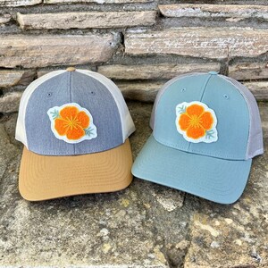 May include: Two trucker hats with a flower embroidered on the front. One hat is gray with a brown brim and the other is blue with a gray brim.