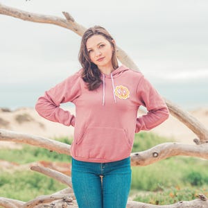 May include: A woman wearing a pink hooded sweatshirt with a yellow and pink monogram patch on the left chest. She is standing in front of a wooden branch.