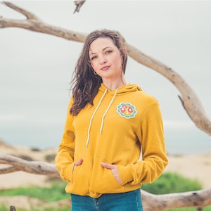 May include: A woman wearing a yellow hoodie with a peace sign patch on the left side. The patch is a colorful design with the letters "SO" inside the peace sign.