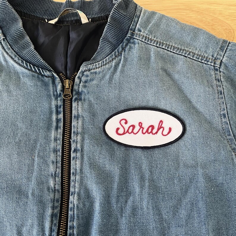May include: A blue denim jacket with a white oval patch that says "Sarah" in red cursive lettering.
