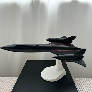 Pode incluir: Um modelo de avi&atilde;o SR-71 Blackbird preto com linhas de destaque vermelhas, montado em um suporte branco. O modelo &eacute; exibido em uma superf&iacute;cie preta contra um fundo branco. A aeronave tem um design elegante e aerodin&acirc;mico.