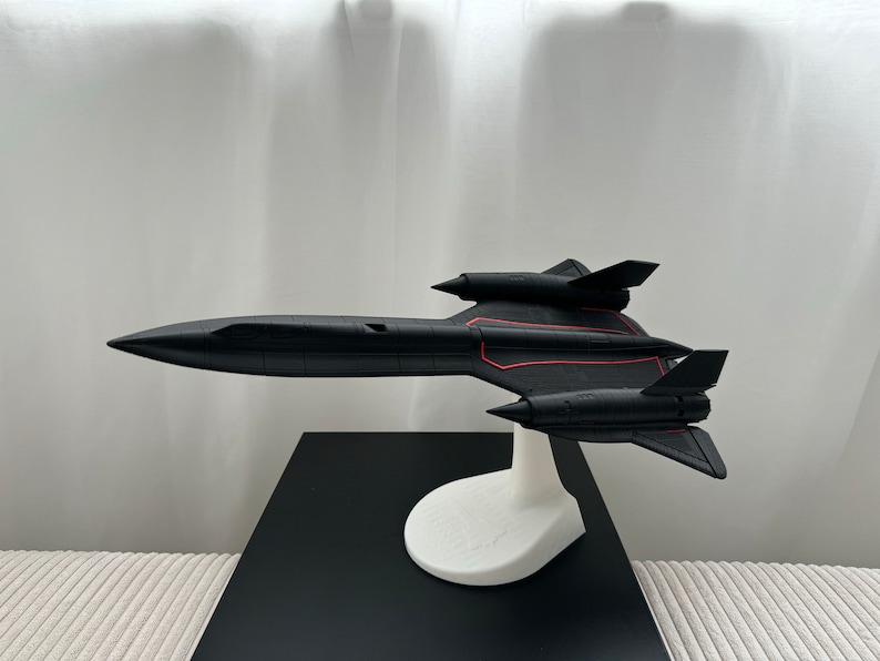 Pode incluir: Um modelo de avi&atilde;o SR-71 Blackbird preto com linhas de destaque vermelhas, montado em um suporte branco. O modelo &eacute; exibido em uma superf&iacute;cie preta contra um fundo branco. A aeronave tem um design elegante e aerodin&acirc;mico.