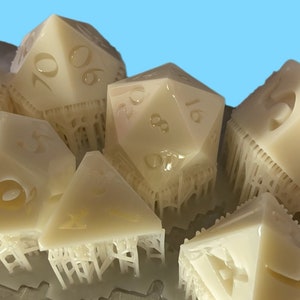 Pode incluir: Um conjunto de dados de v&aacute;rias faces em branco sujo para jogos de tabuleiro. Os dados t&ecirc;m v&aacute;rias formas geom&eacute;tricas, com n&uacute;meros gravados nas suas superf&iacute;cies. Parecem rec&eacute;m-sa&iacute;dos de uma impressora 3D, com estruturas de suporte ainda presas.
